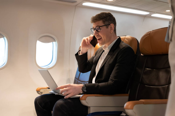Νεαρός ελκυστικός επιχειρηματίας με κοστούμι με γυαλιά που μιλάει στο κινητό τηλέφωνο και εργάζεται σε φορητό υπολογιστή ενώ κάθεται στην καμπίνα του αεροπλάνου. Έννοια εργασίας και ταξιδιού. - Φωτογραφία, εικόνα