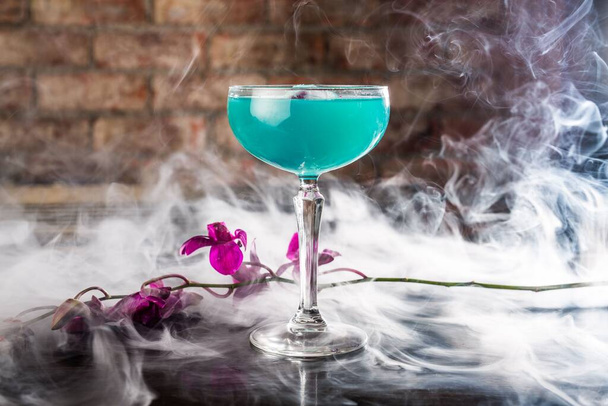 A closeup of a blue cocktail on a table decorated with a purple flower in smoke - Foto, Bild