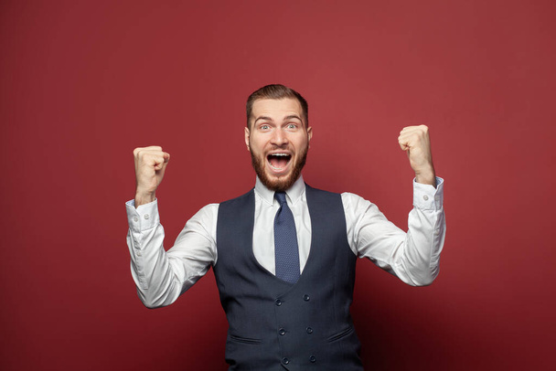 Happy excited businessman portrait. Guy winning, achieve prize or goal, have great news, fist pump and smiling on red banner background - Photo, Image
