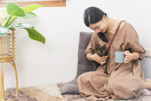 Kisállat szerető koncepció, fiatal ázsiai nő pihenni ivás kávé és játszik macska a nappaliban. - Fotó, kép