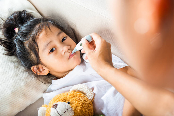 Sick kid. Mother parent checking temperature of her sick daughter with digital thermometer in mouth, child laying in bed taking measuring her temperature for fever and illness, healthcare - Photo, image