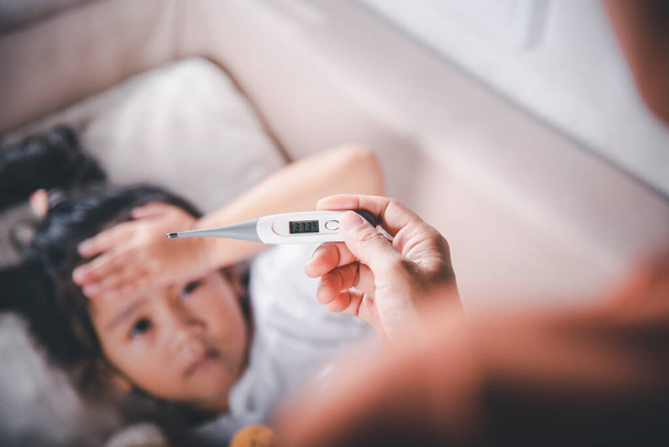 Sick kid. Mother parent checking temperature of her sick daughter with digital thermometer in mouth, child laying in bed taking measuring her temperature for fever and illness, healthcare - Fotó, kép