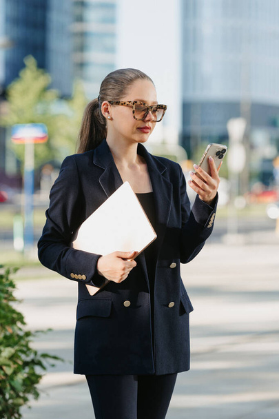A female realtor in a navy blue blazer is holding a laptop and using her smartphone between the office buildings in the financial district. A businesswoman in a cluster of tall buildings. - Photo, image