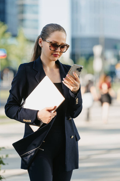 A female realtor in a navy blue blazer is holding a laptop between the office buildings in the financial district. A businesswoman is using her smartphone in a cluster of tall buildings. - Фото, изображение