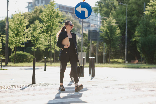 Una agente inmobiliaria femenina en una chaqueta azul marino sostiene una computadora portátil mientras cruza la calle en el centro de la ciudad. Una mujer de negocios está paseando cerca de la línea de árboles. - Foto, imagen