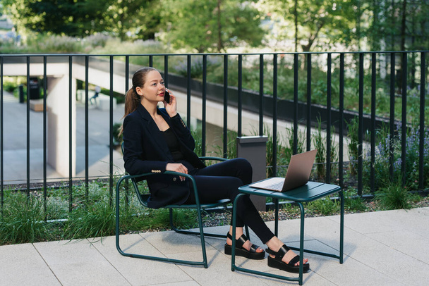 Μια γυναίκα μεσίτρια με σακάκι κάνει μια συζήτηση σε ένα κινητό τηλέφωνο σε ένα σύγχρονο αστικό χώρο στην οικονομική περιοχή. Μια επιχειρηματίας κάθεται στο τραπέζι σε μια συστάδα από ψηλά κτίρια. - Φωτογραφία, εικόνα