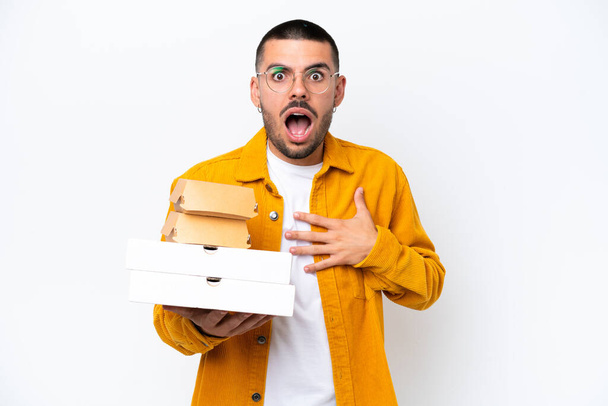 Young caucasian man holding pizzas and burgers isolated on background surprised and shocked while looking right - Photo, Image