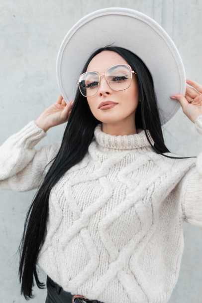 Modieus mooi meisje met een stijlvolle bril in een vintage gebreide trui draagt een hoed in de buurt van een grijze muur op straat en kijkt naar de camera - Foto, afbeelding