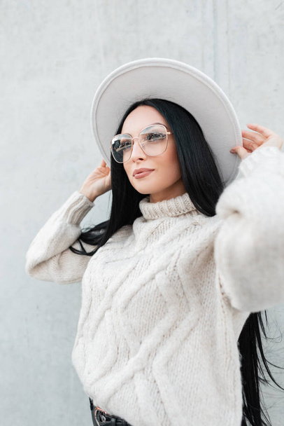 Portrait of trendy pretty woman with fashion glasses in knitted sweater and hat on the street near a gray wall. Female urban autumn style look - Photo, image