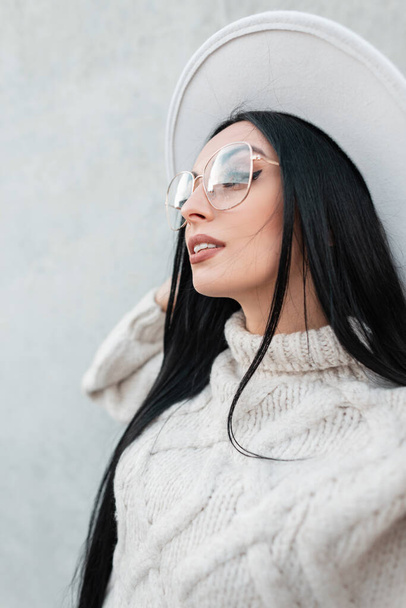 Urban female portrait of beautiful woman model with white hat with vintage glasses in trendy knitted sweater on a gray wall on the street - Photo, image