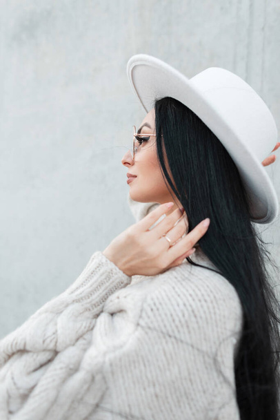 Fashionable beautiful young woman with long brunette hair in a trendy white vintage sweater with a hat and glasses on a gray concrete background - Photo, image