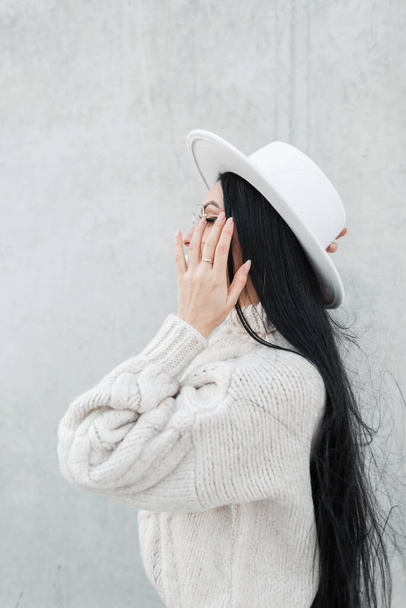 Stylische schöne junge Frau Modell mit langen schwarzen Haaren trägt modische Kleidung mit einem Vintage-Strickpullover und einem Hut in der Nähe einer grauen Betonwand außerhalb. Frauenprofil - Foto, Bild