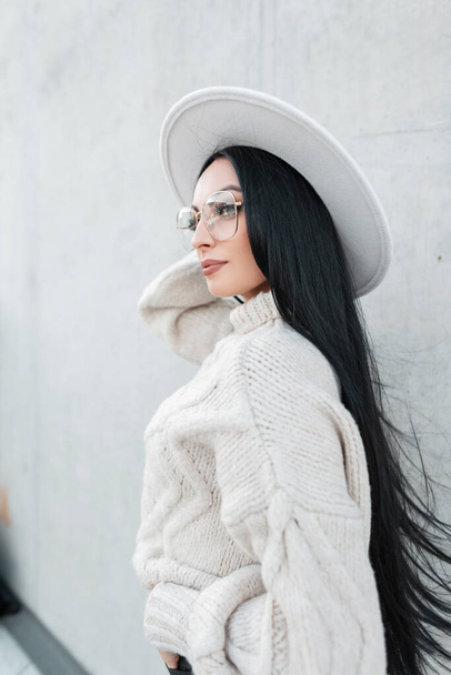 Κομψό όμορφη νεαρή γυναίκα με μακριά καστανά μαλλιά και μοντέρνα γυαλιά σε ένα μοντέρνο λευκό καπέλο με πλεκτό vintage πουλόβερ βόλτες στο δρόμο κοντά στο τσιμεντένιο τοίχο - Φωτογραφία, εικόνα