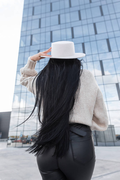 Beautiful young sexy girl with healthy long brunette hair in fashionable clothes with a vintage knit sweater, hat and leather pants walking in the city, back view - Photo, Image