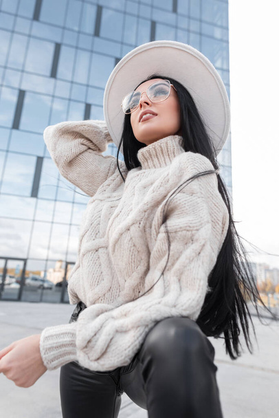 Fashionable beautiful brunette model with glasses in a vintage knit sweater, hat, and leather pants poses near a glass modern building - 写真・画像