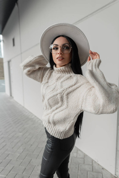 Divatos városi lány modell szemüveges divatos kötött pulóver és kalap séták az utcán, közel egy szürke modern épület - Fotó, kép