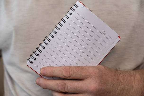 A middle-aged man is holding an open notebook on a spring. The blank pages of the notebook are in a line. Notebook in a man's hand. Middle section of a mature man in a gray T-shirt. Selective Focus. - Foto, Imagen
