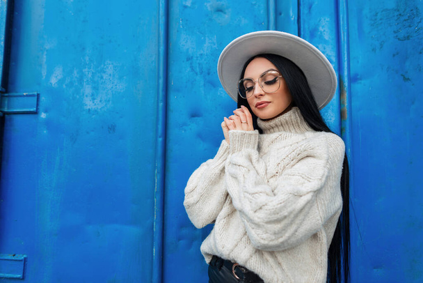 Мода Кавказская женщина модель с винтажными очками в модном трикотажном свитере и белой шляпе стоит возле синей гранж-металлической стены - Фото, изображение