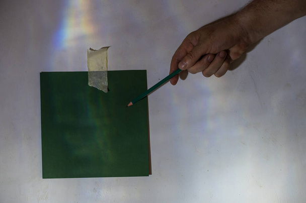 Olgun bir adamın eli kalemle bir kağıt parçasına işaret eder. Duvara sabitlenmiş boş yeşil kare kağıt parçası. Gri alçılı bir duvara soyut bir ışık ve gölge çizimi. Seçici odaklanma. Alçak tuş - Fotoğraf, Görsel