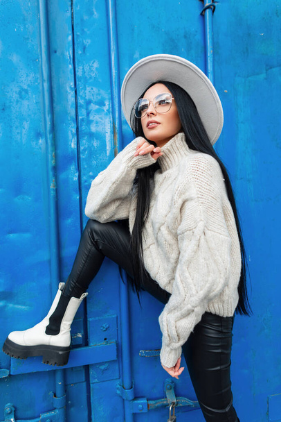 Stylish beauty fashion girl model with trendy glasses and hat in white knit fashion sweater, leather pants and boots poses near a color blue metal wall - Fotografie, Obrázek