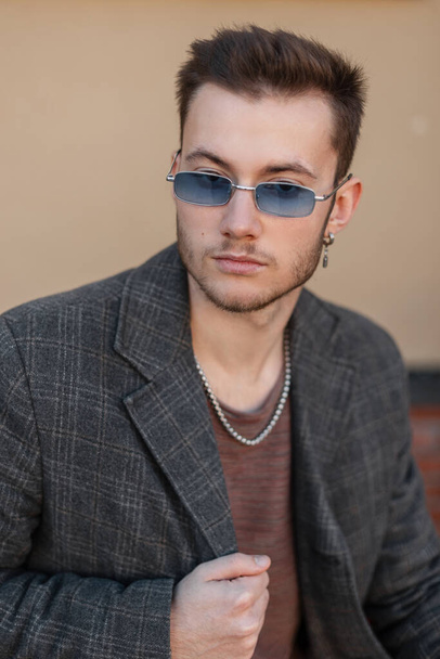 Fashionable young guy hipster with blue sunglasses in trendy coat sits in the city - Photo, image