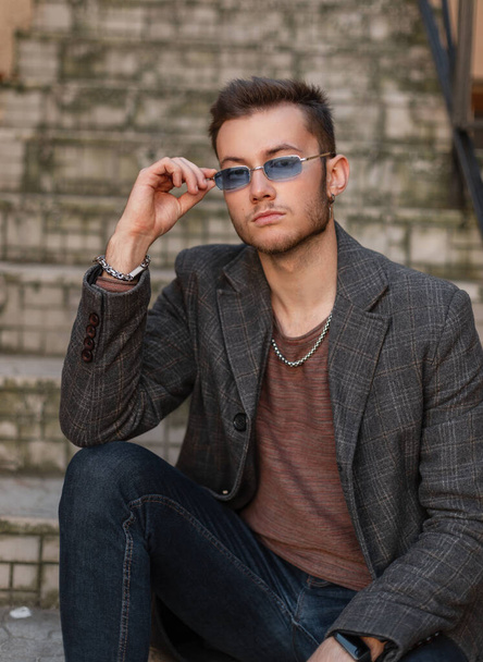 Fashionable hipster guy in a stylish casual coat, T-shirt and jeans sits on the vintage steps and puts on his stylish blue sunglasses - Photo, image