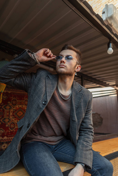 Όμορφος hipster μοντέλο άνθρωπος με μοντέρνο παλτό, φούτερ και τζιν φοράει ένα μπλε γυαλιά ηλίου και κάθεται στο δρόμο - Φωτογραφία, εικόνα