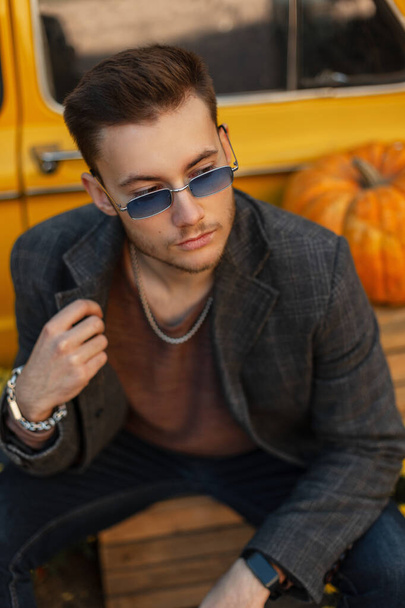 Modaya uygun, genç, yakışıklı iş adamı hippi mavi, klasik güneş gözlüklü, şık montlu, eski sarı, balkabaklı bir arabanın yanında oturuyor. Sonbahar erkek tarzı giysiler - Fotoğraf, Görsel