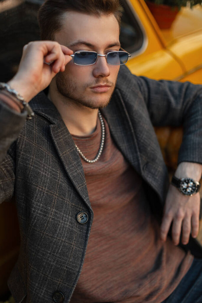 Модный бизнесмен хипстер в повседневной одежде в модном пальто и роскошных часах носит стильные солнцезащитные очки и сидит рядом с желтым ретро-автомобилем - Фото, изображение