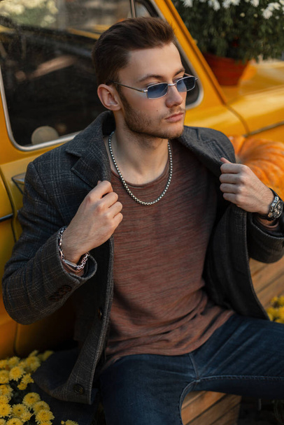 Fashion handsome businessman with sunglasses in stylish coat and fashionable sweatshirt sits near a retro yellow car on the street - Fotografie, Obrázek