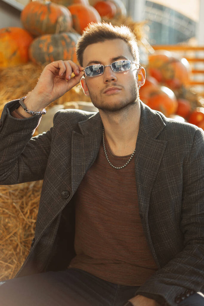 Fashionable fall handsome man with vintage sunglasses in coat and sweatshirt sits near a pumpkins in countryside - 写真・画像