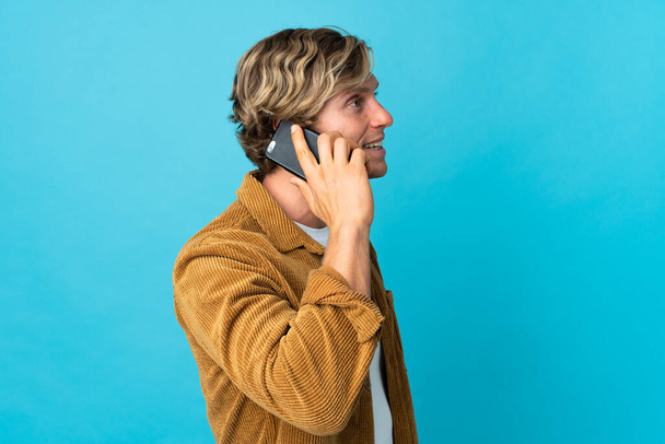 Английский человек на изолированном синем фоне ведет разговор с кем-то по мобильному телефону - Фото, изображение