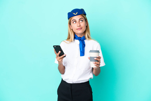 Stewardessa samolotu urugwajska kobieta odizolowana na niebieskim tle trzymająca kawę do zabrania i telefon komórkowy myśląc coś - Zdjęcie, obraz