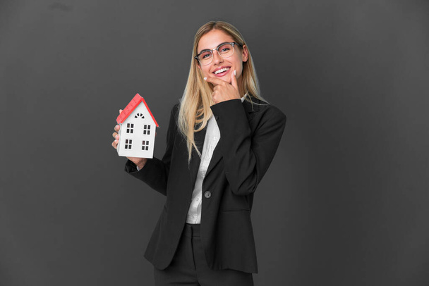 Blonde Uruguayan girl holding a house toy isolated on black background happy and smiling - Foto, Imagem