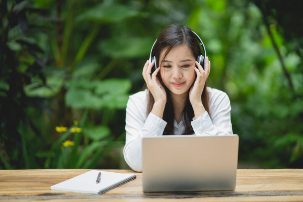 boldog fiatal ázsiai nő mosolyog laptoppal és fehér fejhallgatót visel, miközben a kávézóban ül. Fiatal ázsiai nő ül egy kávézóban és videokonferencia laptopon. - Fotó, kép