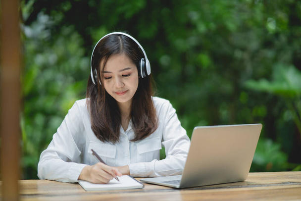 ノートパソコンを使って白いヘッドフォンをつけてノートを書く幸せな若いアジアの女性の笑顔。若いアジア人女性座っていますでコーヒーショップとビデオ通話会議上のラップトップ. - 写真・画像