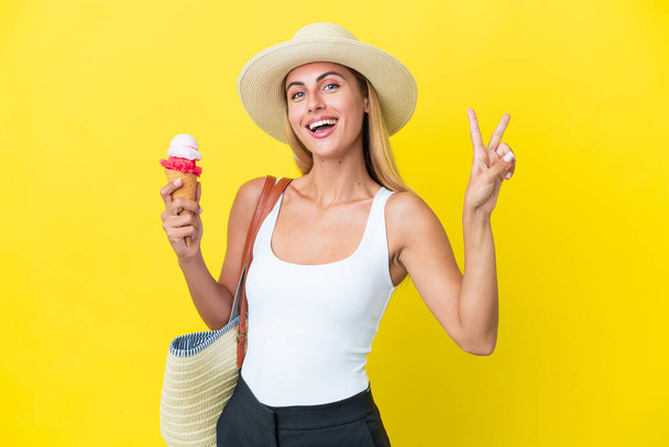 Blondes uruguayisches Mädchen im Sommer mit Eis auf gelbem Hintergrund, lächelnd und mit Siegeszeichen - Foto, Bild