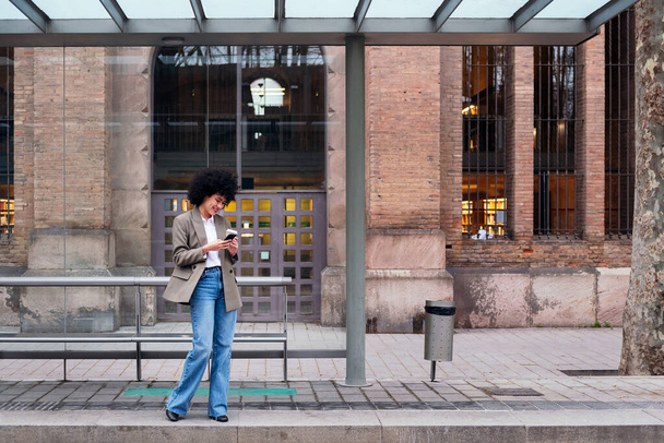 young latin woman waiting at the bus stop checking her mobile phone, concept of urban and modern lifestyle - Fotoğraf, Görsel