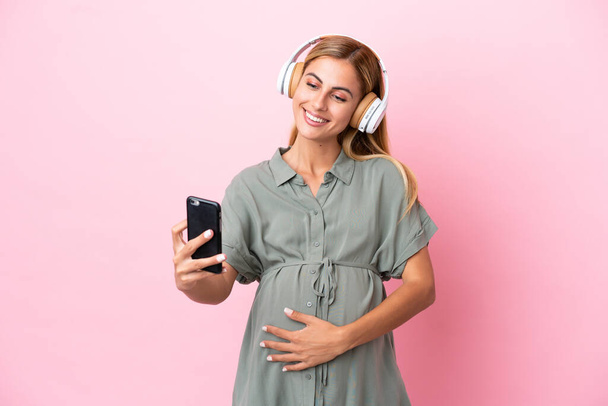 Mujer uruguaya joven aislada sobre fondo azul embarazada y escuchando música con un móvil - Foto, imagen