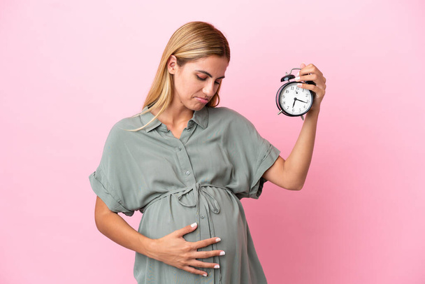 Young Uruguayan woman isolated on blue background pregnant and holding clock with stressed expression - Foto, imagen