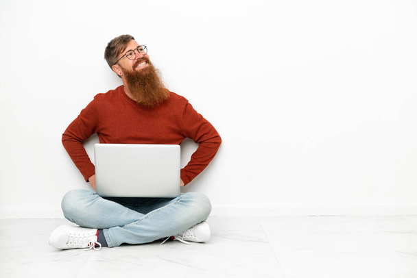 Junge rötliche kaukasische Mann mit Laptop isoliert auf weißem Hintergrund posiert mit Armen an der Hüfte und lächelt - Foto, Bild