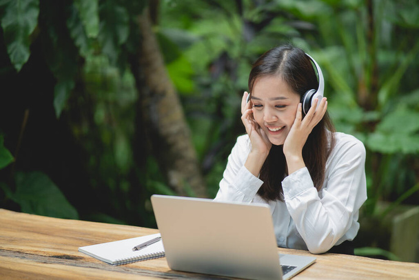 幸せな若いアジアの女性はノートパソコンを使って笑顔で白いヘッドフォンをしてカフェに座っています。若いアジア人女性座っていますでコーヒーショップとビデオ通話会議上のラップトップ. - 写真・画像