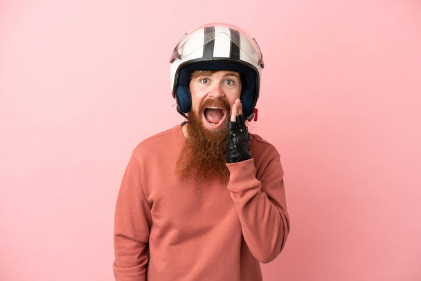 Νεαρός ερυθρός καυκάσιος άνδρας με κράνος μοτοσικλέτας απομονωμένος σε ροζ φόντο με έκπληξη και σοκαρισμένη έκφραση προσώπου - Φωτογραφία, εικόνα