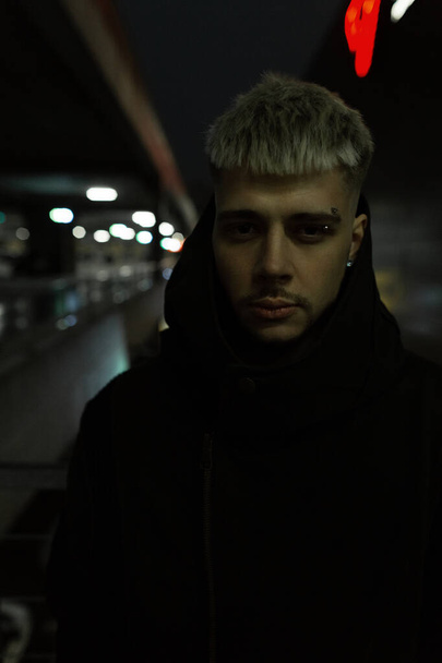 Retrato de noche urbana de moda de un guapo tipo brutal serio en una chaqueta negra con capucha se para en la calle por la noche - Foto, imagen