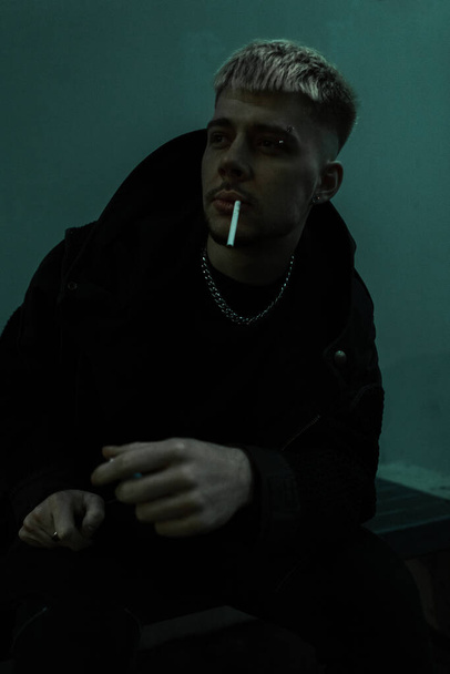 Cool brutální muž v černém oblečení sedí venku a kouří cigaretu v nočním městě - Fotografie, Obrázek
