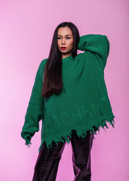 Fashionable stylish beautiful young Vietnamese girl with hair in a trendy ripped green sweater with black leather pants poses on a pink background - Foto, immagini