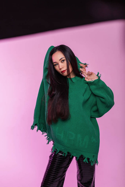 Stylová krásná mladá korejská dívka se zdravými dlouhými vlasy v trendy trhaný zelený svetr s koženými kalhotami ve studiu s růžovými světly - Fotografie, Obrázek