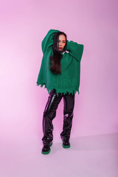 Beautiful young Asian model girl in fashionable clothes with green sweater and leather pants with boots in the studio on a pink background with pink light - Photo, Image