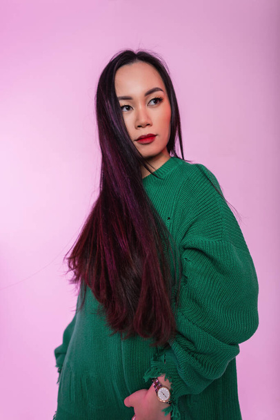 Chica asiática hermosa de moda con cabello largo saludable en un suéter de punto desgarrado verde de moda en un estudio con luz rosa - Foto, imagen