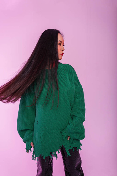 Красива молода стильна модель жінки зі здоровим волоссям в модному зеленому в'язаному светрі зі шкіряними штанами на зеленому тлі в студії
 - Фото, зображення